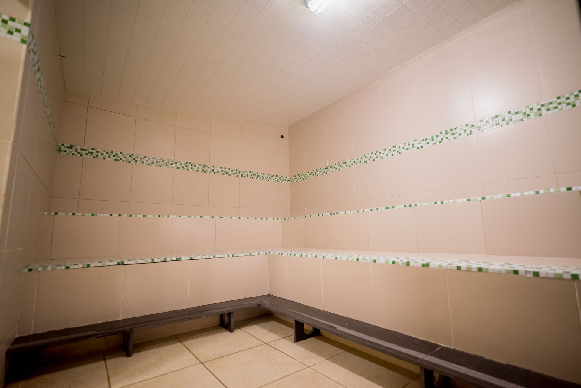Banheiro vazio com paredes revestidas de azulejos, bancos e prateleiras. Tons de bege claro com detalhes em verde.