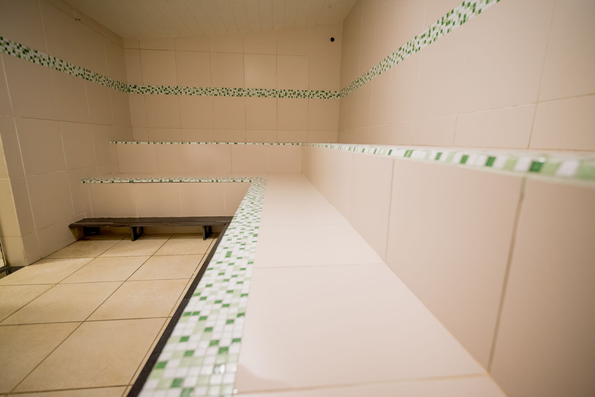 Sala de vapor revestida com azulejos e bancos em níveis, com detalhes em mosaico verde.