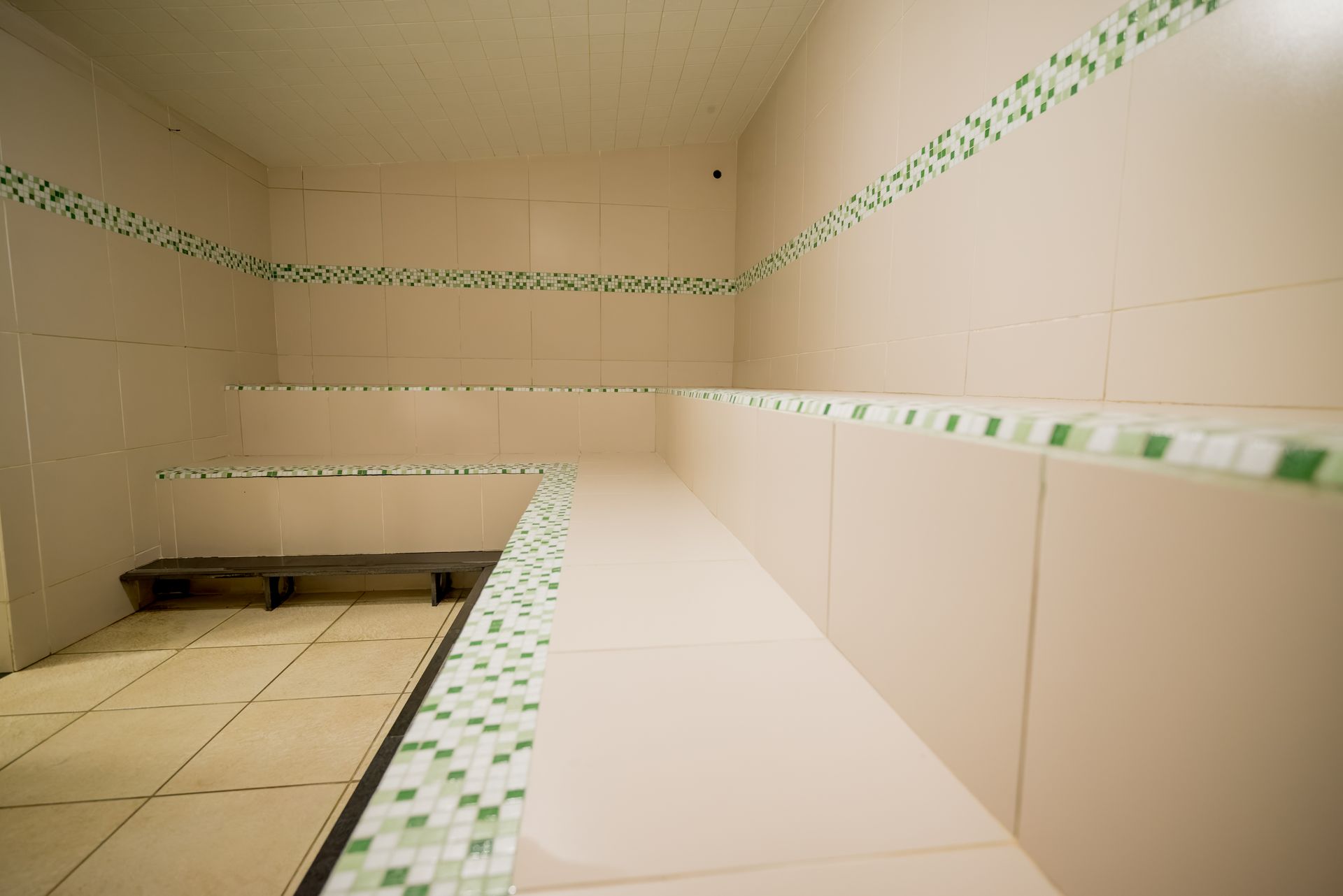 Interior de uma sala de vapor com paredes revestidas de azulejos e bancos. Azulejos bege claro com detalhes em mosaico verde e banco de madeira.