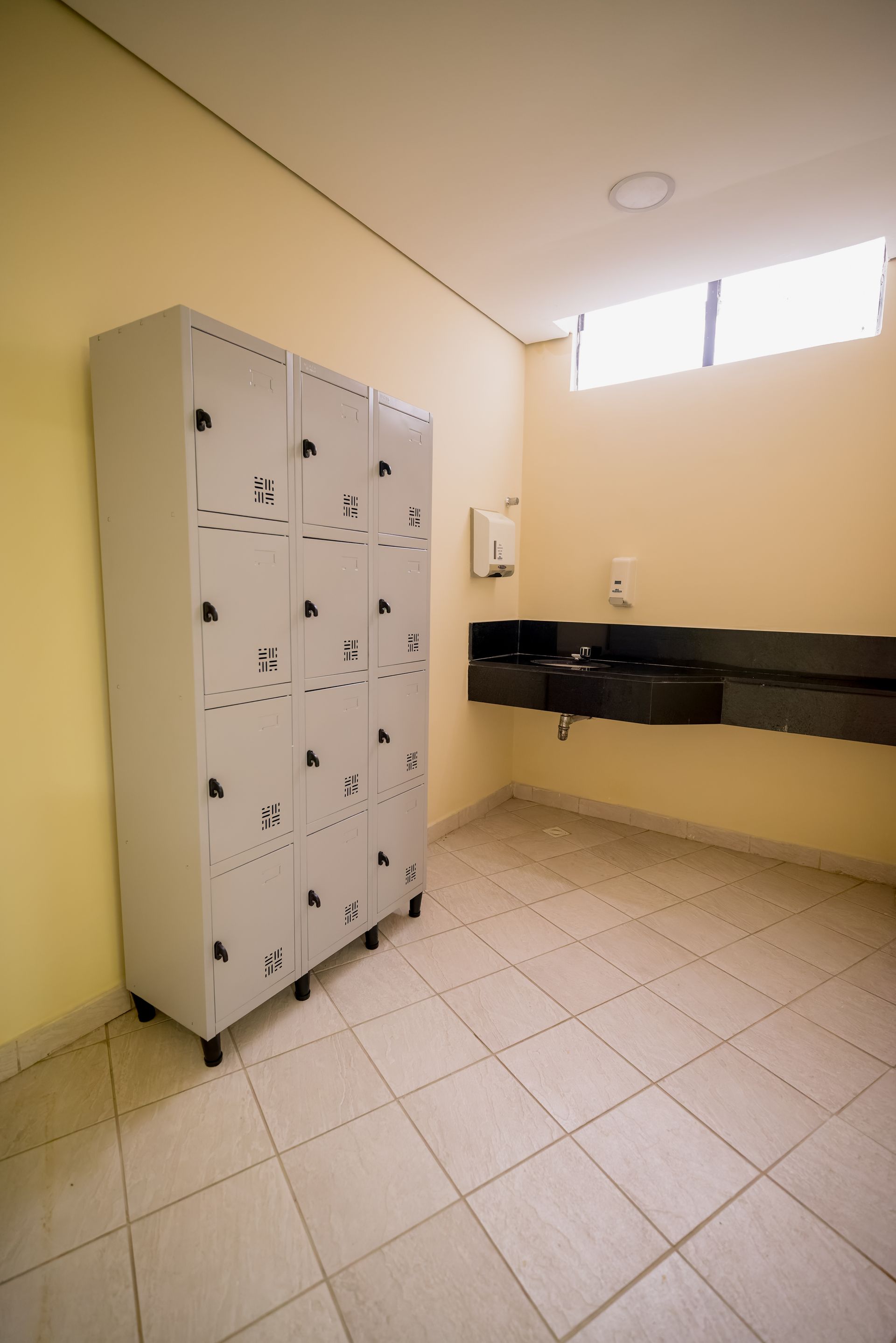 Vestiário com paredes bege, armários, pia e piso de azulejo. Um dispensador de sabonete está instalado na parede.
