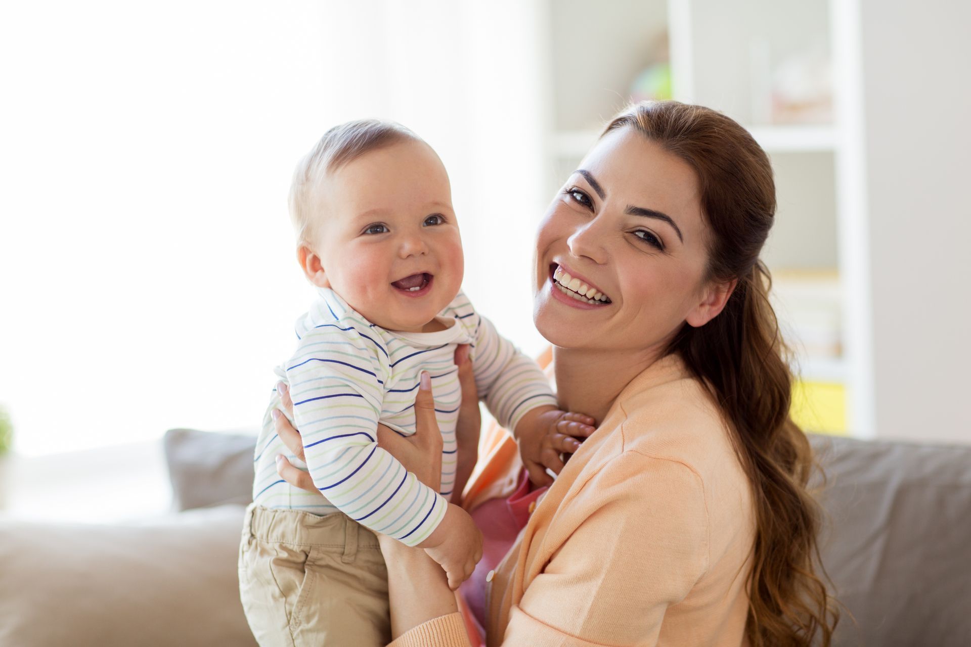 Woman holding and smiling at a laughing baby indoors; both are facing the camera.