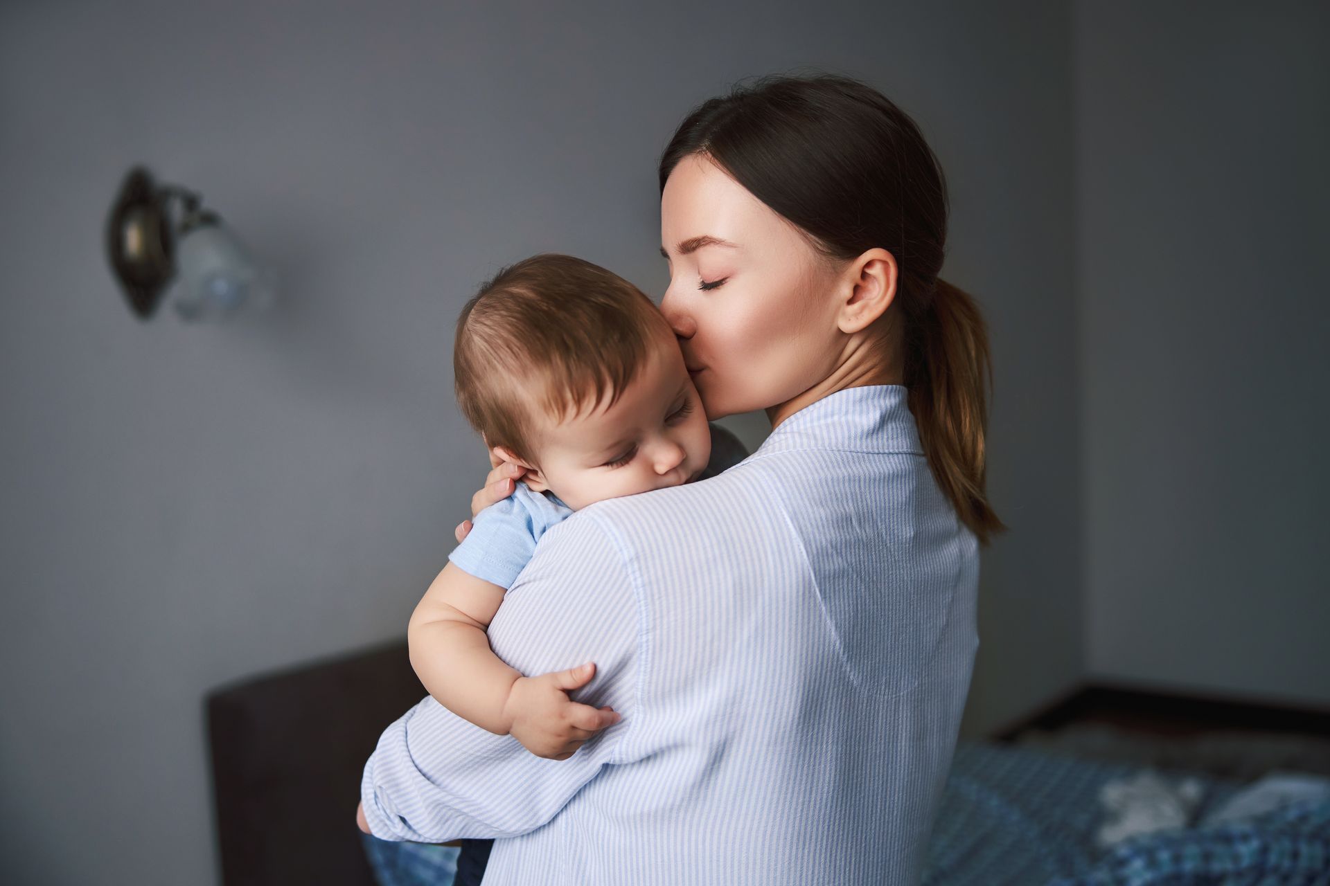 Woman holding sleeping baby, kissing its head indoors.