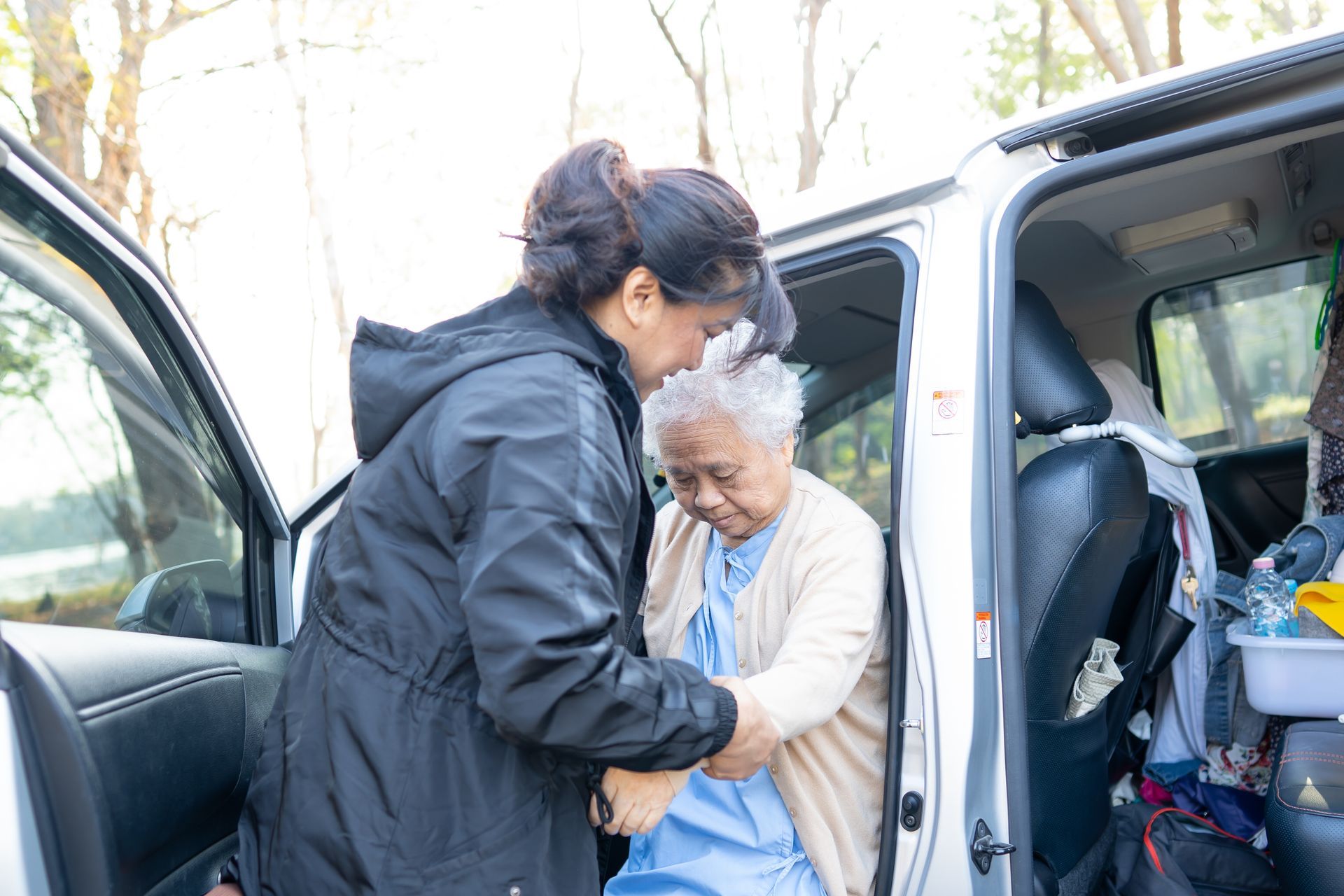 Woman assisting an elderly person out of a car. Outdoors, trees in background.