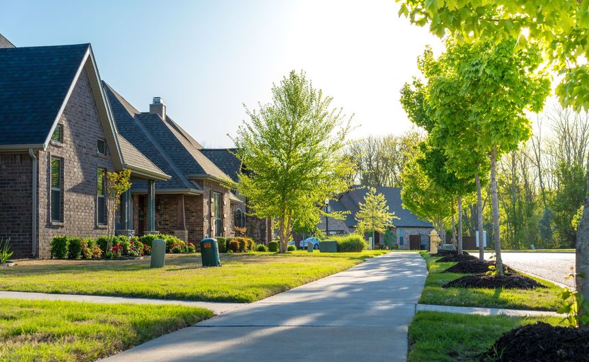 Suburban street with brick houses, green trees, and a sunlit sidewalk on a clear day