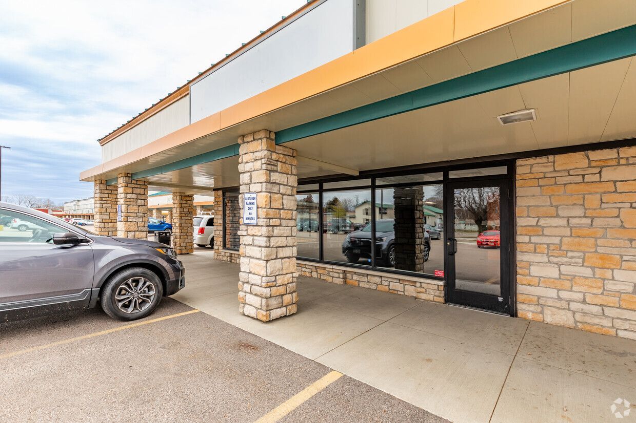 Exterior view of a commercial building with a gray car parked in front. Stone columns, glass windows, and a tan awning are visible.