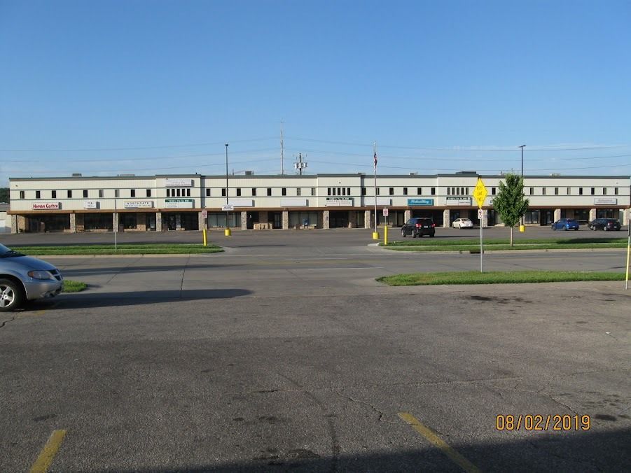 Strip mall with a long, flat facade and multiple storefronts. A parking lot in the foreground with a car.