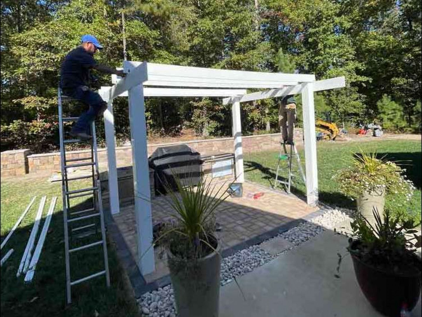 Person on a ladder installing a white pergola in a garden patio area.