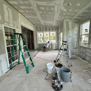 Interior room under renovation with drywall patches, ladders, tools, and a worker near the windows