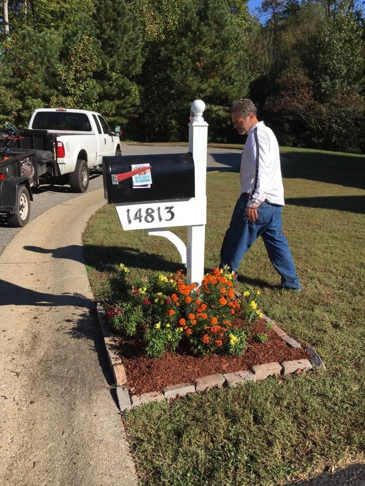 Man placing a white mailbox labeled 14813 beside a flower bed and driveway on a sunny street.