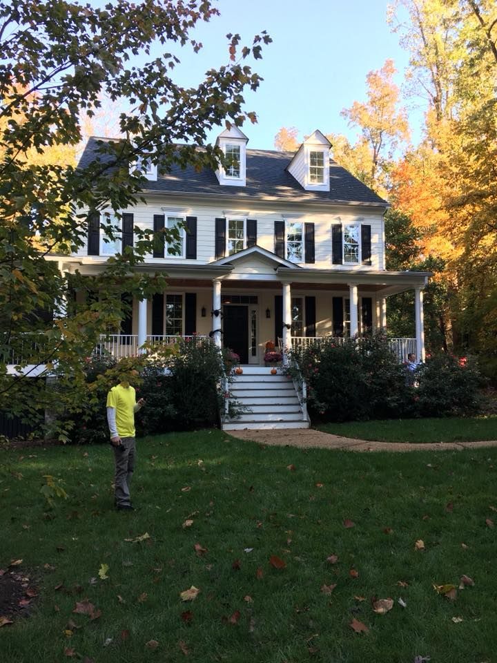 Large white house with a wraparound porch, surrounded by trees; a person in a yellow jacket stands on the lawn.