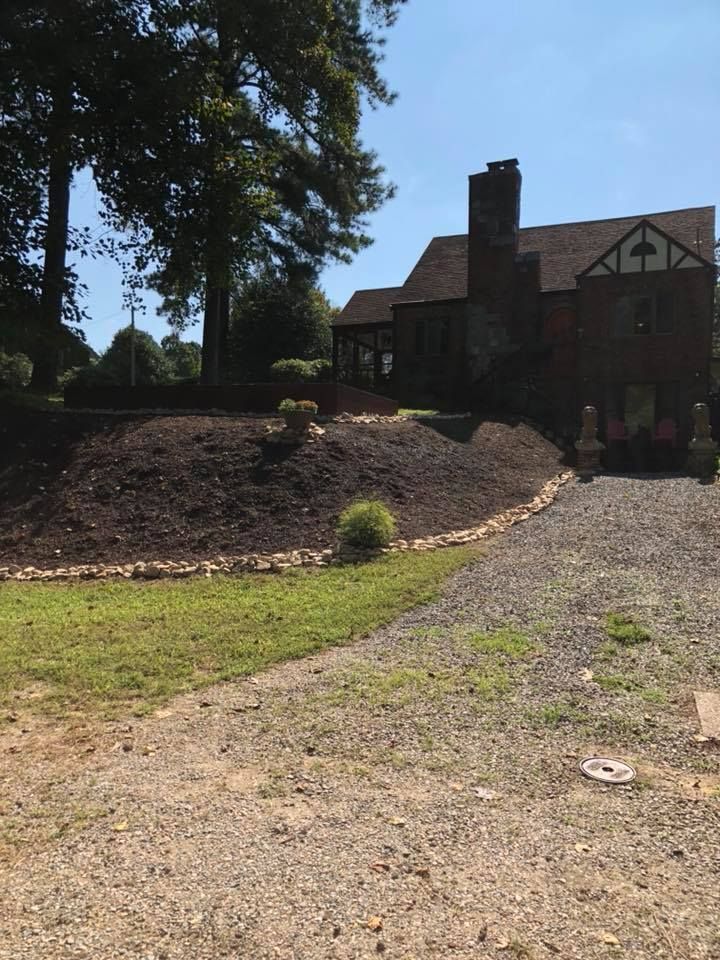 Brick house with a gravel driveway, mulched garden bed, and large trees in bright sunlight