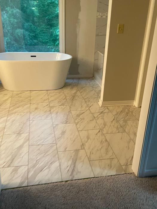 Bathroom with white freestanding tub, marble tile floor, and large window view of greenery.