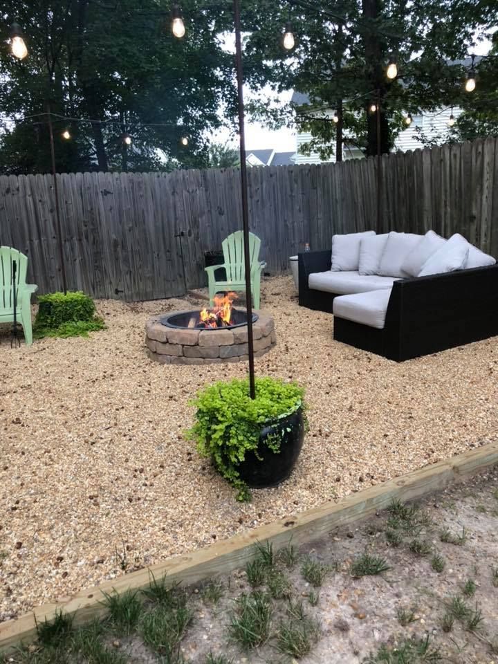 Backyard patio with gravel, a fire pit, black sectional sofa, and green chairs under string lights