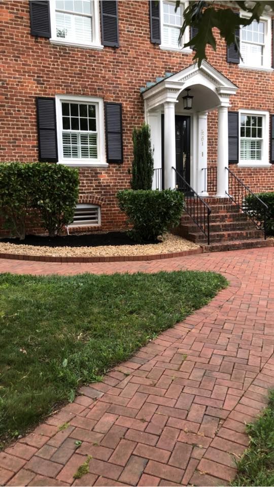 Brick house entrance with white columns, red brick steps, and a curved brick walkway through a green lawn.