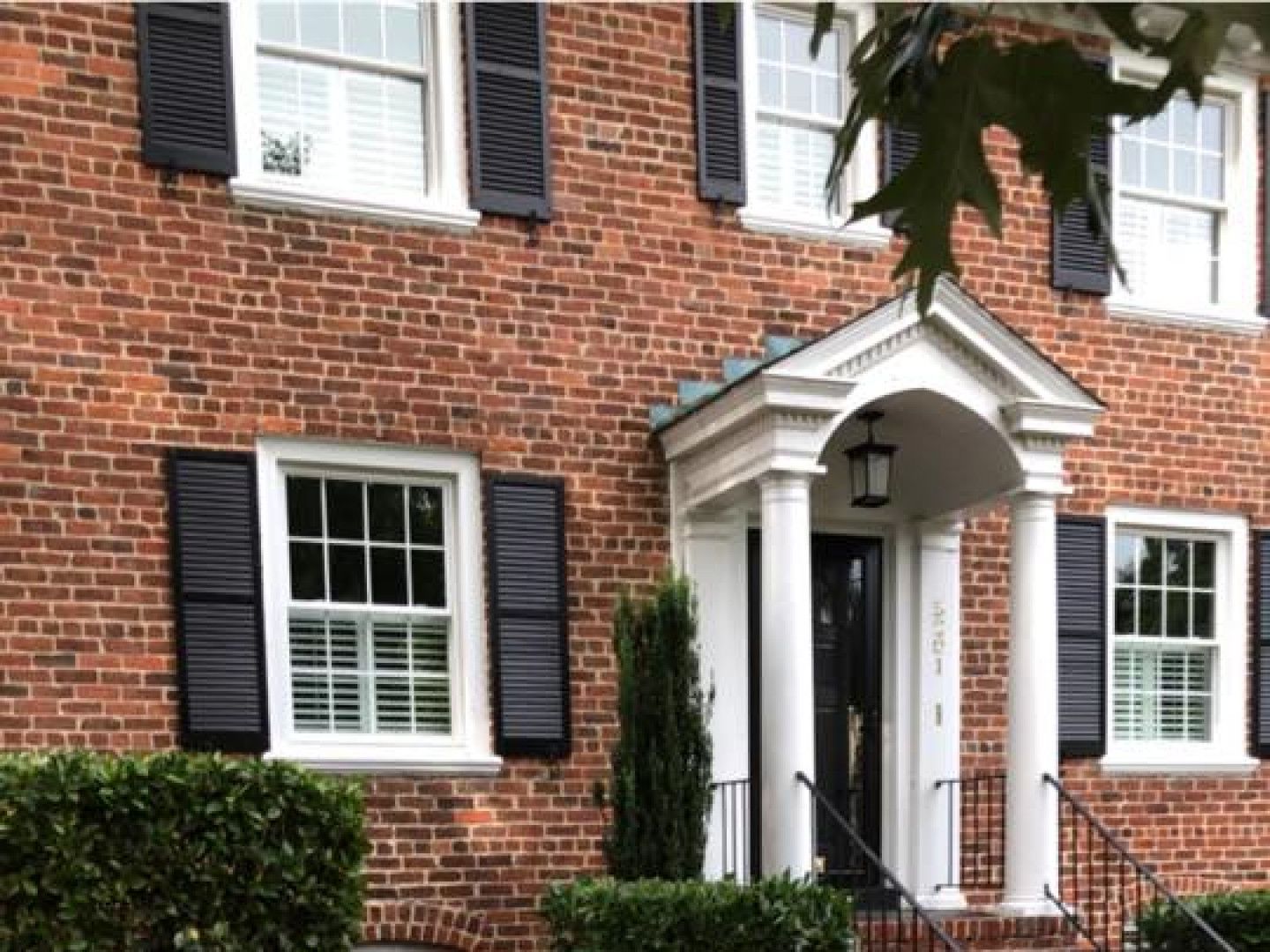 Brick house entrance with white columns, black shutters, and a front door under a small porch.