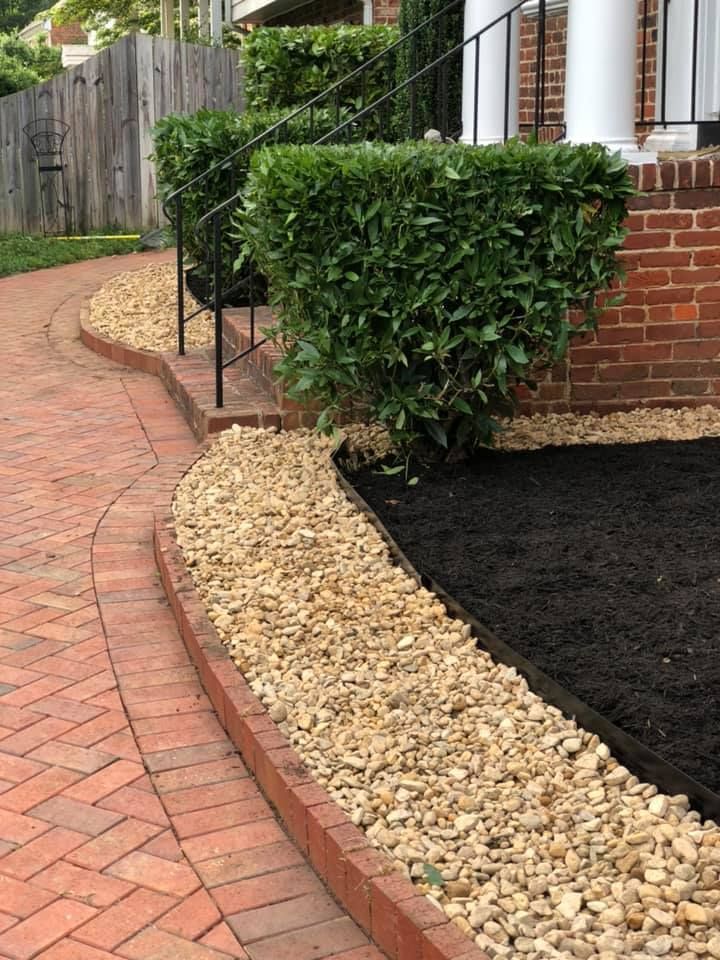 Curved brick walkway beside gravel edging, trimmed hedge, and brick house front yard