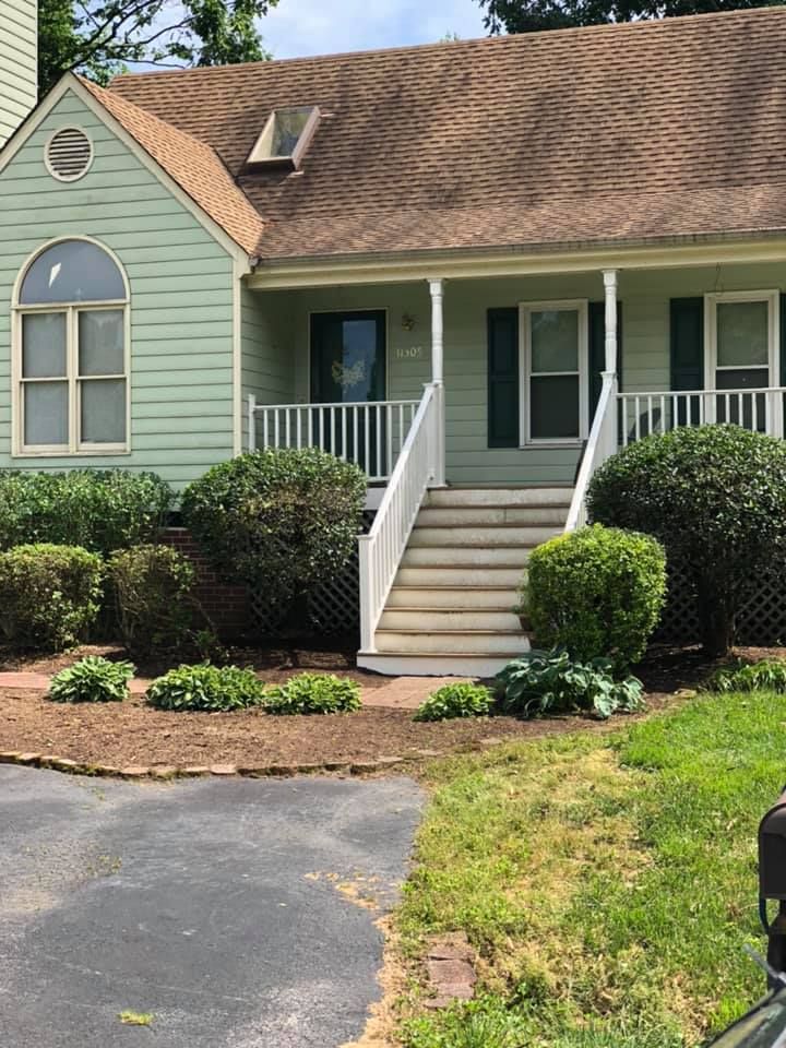 Front entrance of a light green house with white trim, porch steps, shrubs, and a driveway.