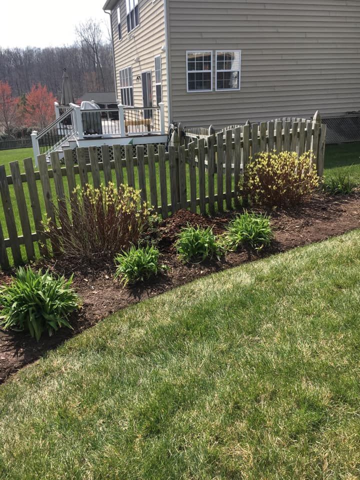 Backyard flower bed with green shrubs along a wooden fence beside a beige house.