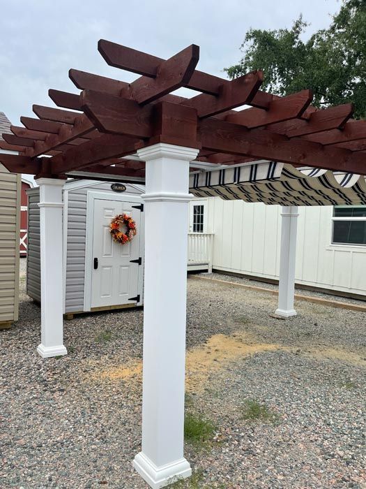 White pergola with dark wood beams over a gravel yard beside a shed and fence