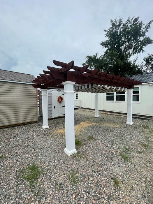 White pergola with maroon slats beside a beige building on a gravel lot under a cloudy sky