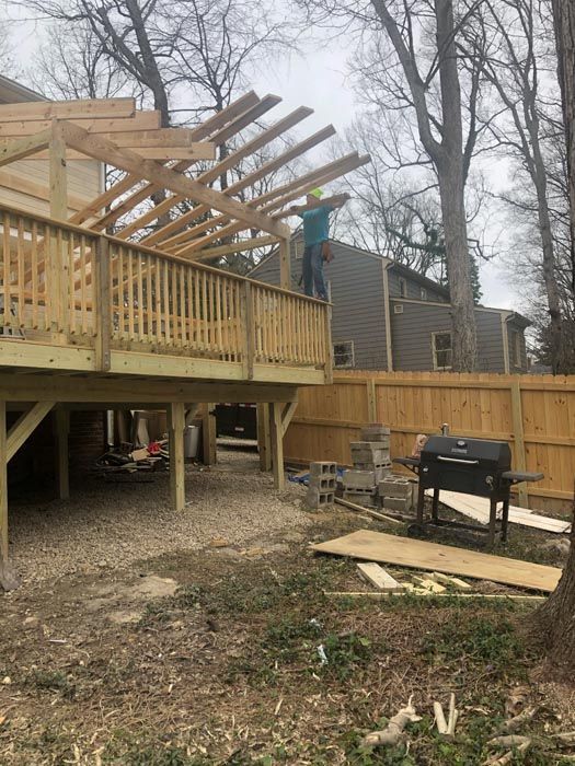 Worker on a wooden deck roof frame beside a house, with unfinished beams and outdoor grill below.