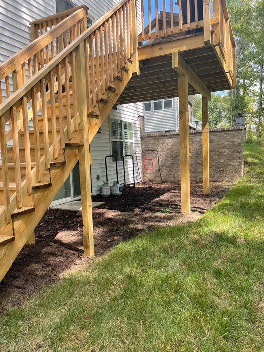 Wooden outdoor deck staircase beside a house, with railing and support posts over a grassy yard.