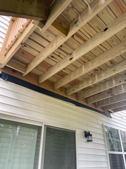 Wooden deck underside with exposed beams above a white house exterior and window