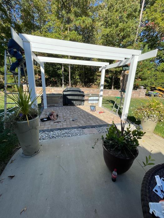 White pergola over a stone patio with potted plants and trees in a sunny backyard
