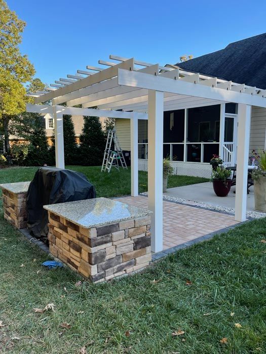 Backyard pergola with white posts, stone bench, and covered grill beside a house
