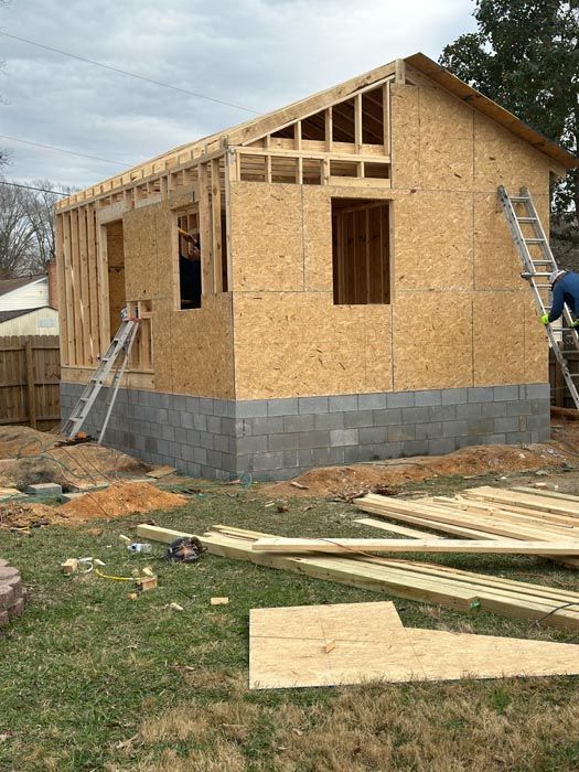 Wood-frame house under construction with exposed plywood walls, ladder, and scattered lumber in the yard