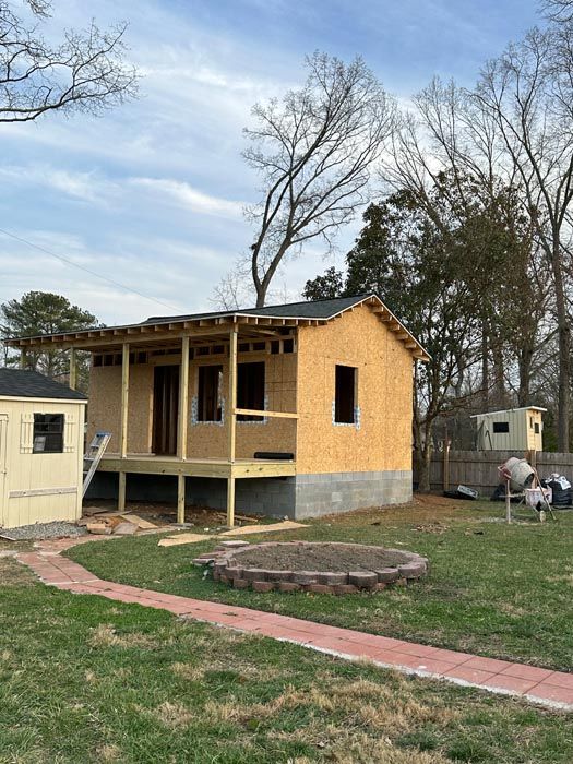 Small yellow house under construction with porch, windows, and a yard with a brick walkway and fire pit.