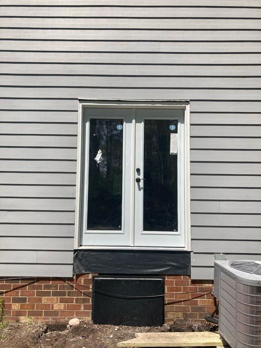 White French doors in a gray siding wall above a brick foundation, with a small black step below.
