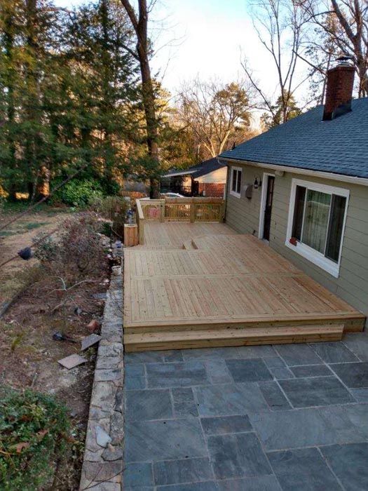Backyard deck under construction beside a house, with a stone patio and wooded yard.