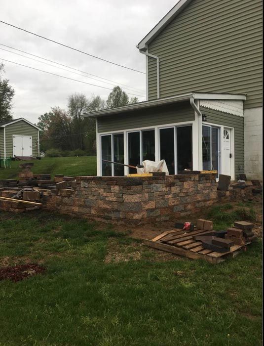 Backyard stone patio under construction beside a house, with stacked pavers and screened porch.