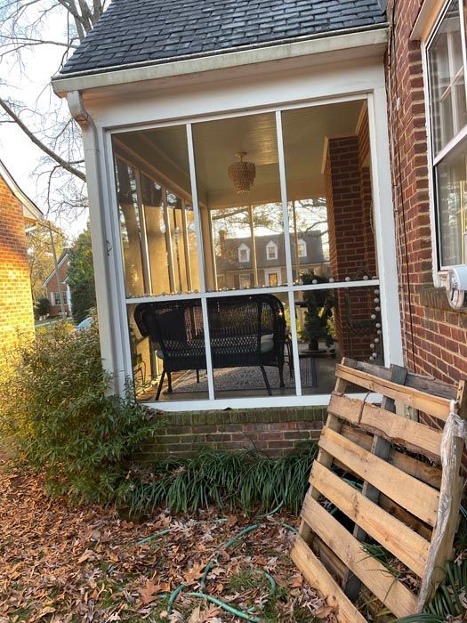 Covered front porch with chair and brick house, framed by leaf-covered yard and stacked wooden pallets.