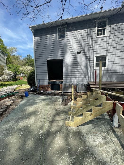 Backyard patio under construction beside gray house, with wooden steps and gravel driveway