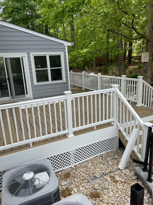White-railed wooden deck beside gray house, with outdoor AC unit and trees in the background.