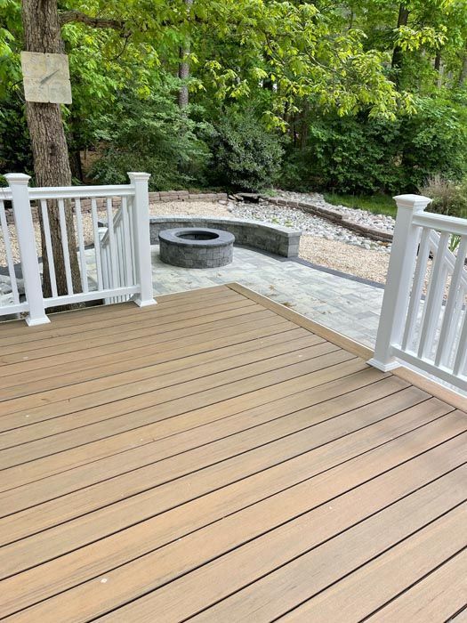 Wooden deck overlooking a stone patio with a built-in fire pit and lush green trees.