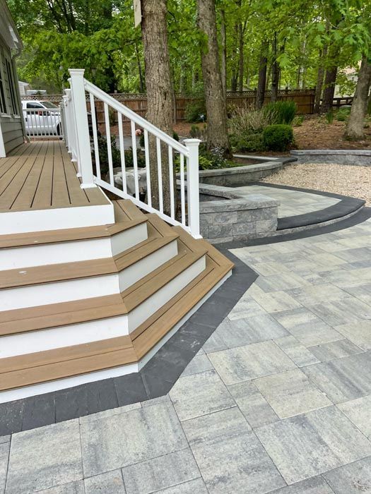 Wooden deck stairs with white railing leading to a gray paver patio in a wooded yard
