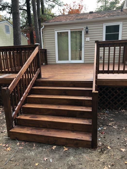 Wooden deck with stairs leading to a beige house, flanked by dark railings and autumn trees.