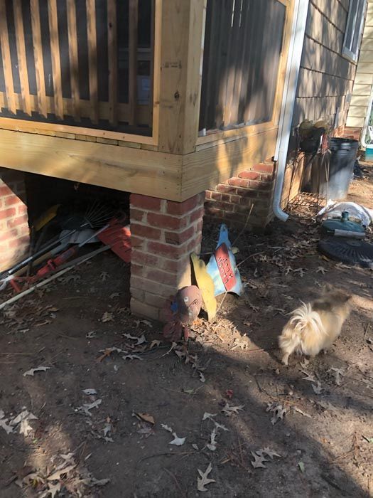 Raised house corner on brick piers, with tools and a small dog on dirt beside the foundation.