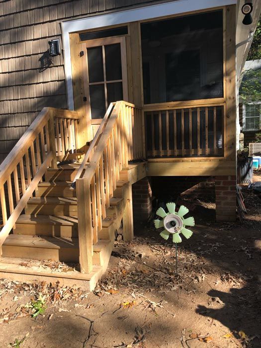 Front porch with wooden stairs and railings leading to a screened entry, beside a green flower decoration in the yard.