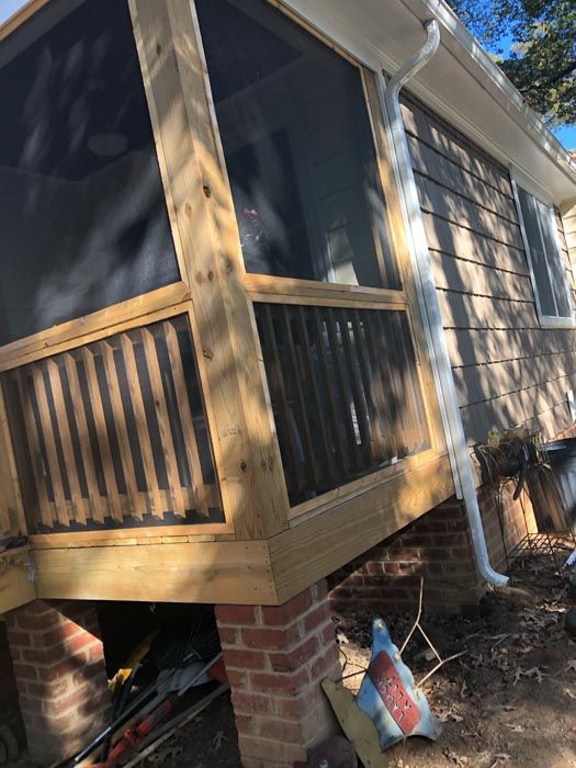 Screened porch with wood railings and brick foundation beside a beige house, viewed at an angle.