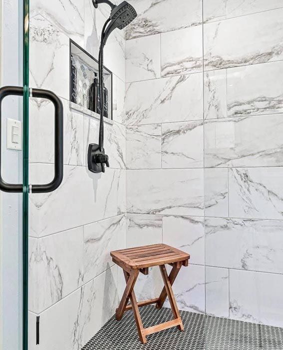 Modern tiled shower with black showerhead and a small wooden stool inside.