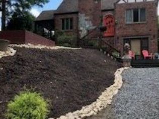 Front yard with dark mulch slope, stone border, and brick house with red door and patio chairs.