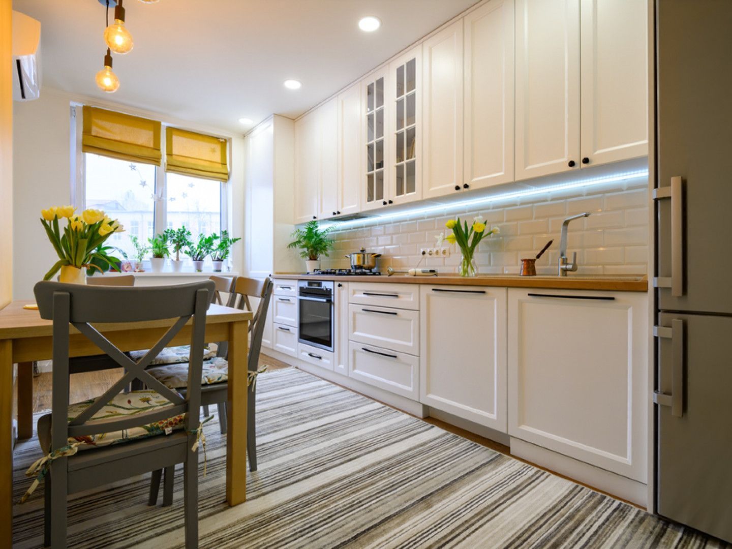 Bright modern kitchen with white cabinets, wooden counters, striped rug, and dining table by the window