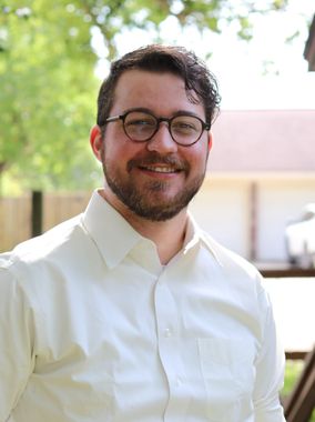 Smiling person with glasses and short beard, wearing a light blue shirt, seated against a plain wall