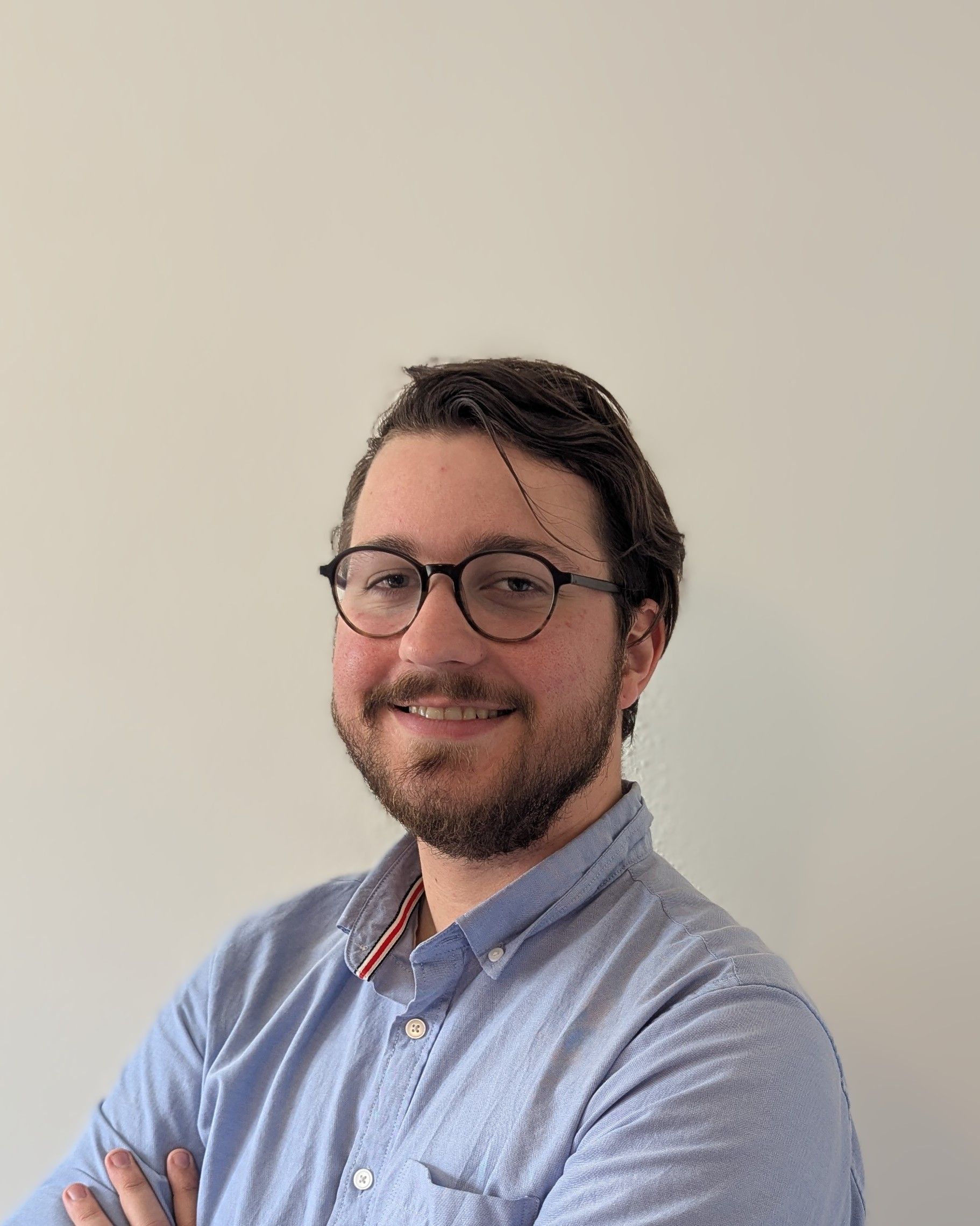 Smiling person with glasses and short beard, wearing a light blue shirt, seated against a plain wall