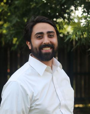 Smiling person with dark hair and beard wearing a light blue button-up shirt against a plain beige wall