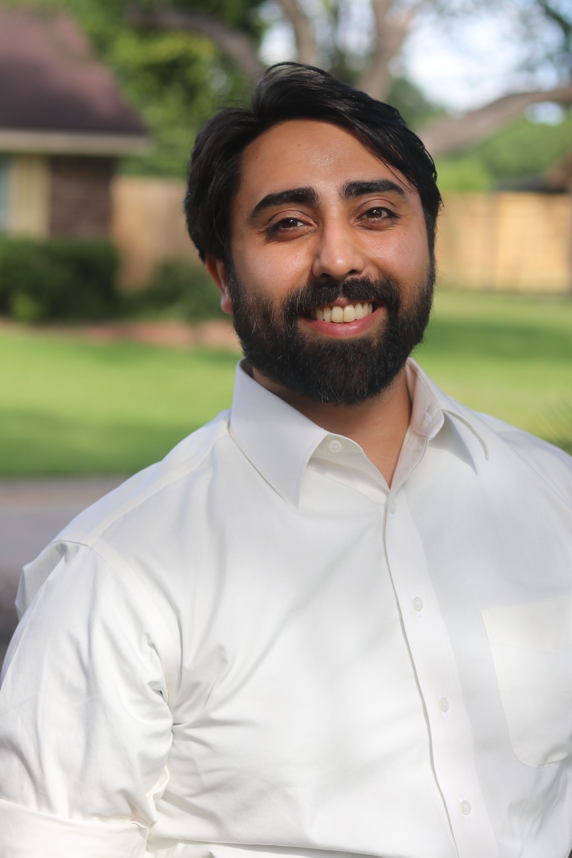 Smiling man in a white shirt standing outdoors with a blurred green lawn and building behind him
